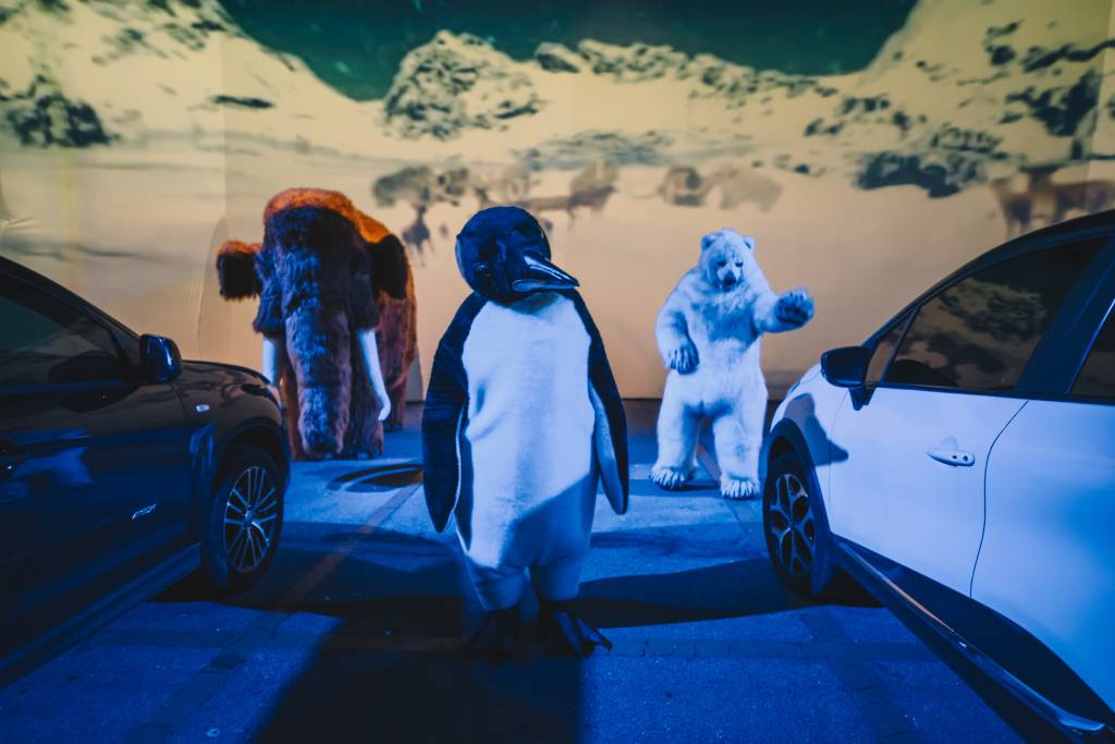 Bonecos de pinguim, mamute e urso polar em frente a dois carros e telão com imagens de neve