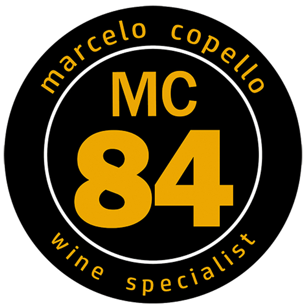 Círculo preto em fundo branco com escritos em amarelo. Na parte superior, lê-se Marcelo Copello em caixa baixa, ao centro as iniciais MC seguidas pelo número 84 em destaque abaixo e na parte inferior, "wine specialist". Selo de qualidade e vinhos do crítico Marcelo Copello. 