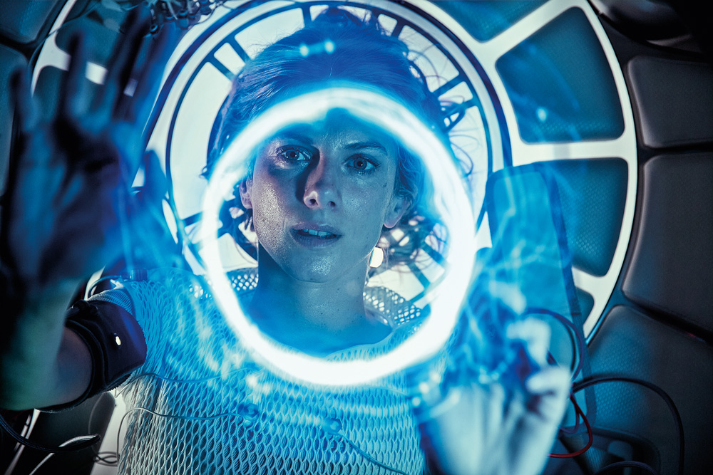 A imagem mostra a atriz em uma câmera criogênica, com um circulo de luz em sua frente e seu rosto mostra uma expressão de dúvida.
