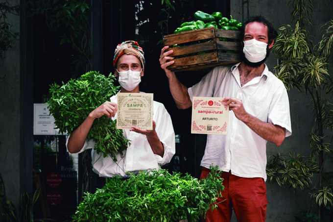 Paola Carosella (à esquerda) e Arpad Spalding (à direita) de máscara segurando legumes e selos da SAMPA+RURAL.