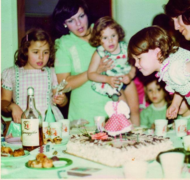Paola Carosella Família reunida envolta de mesa com bolo de aniversário ao centro. Criança segurada por um adulto (à esquerda) para soprar a vela do bolo.