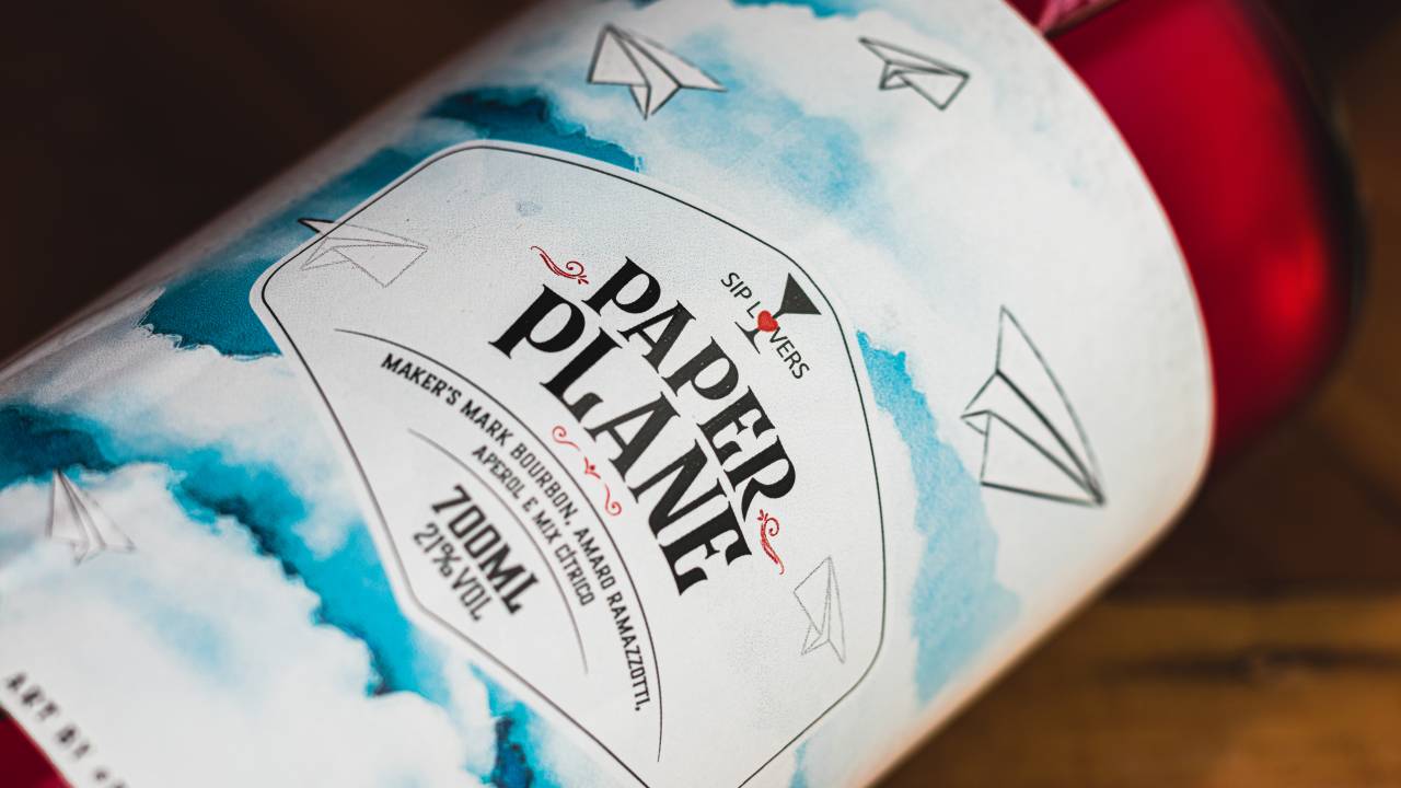 Foto do rótulo do drinque engarrafado 'paper plane' desenhado pelo Eduardo Srur. Fundo branco com nuvens em azul e aviõezinhos de papel.