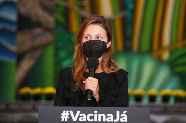 A imagem mostra Patricia Ellen, de máscara, segurando um microfone no Palácio dos Bandeirantes.