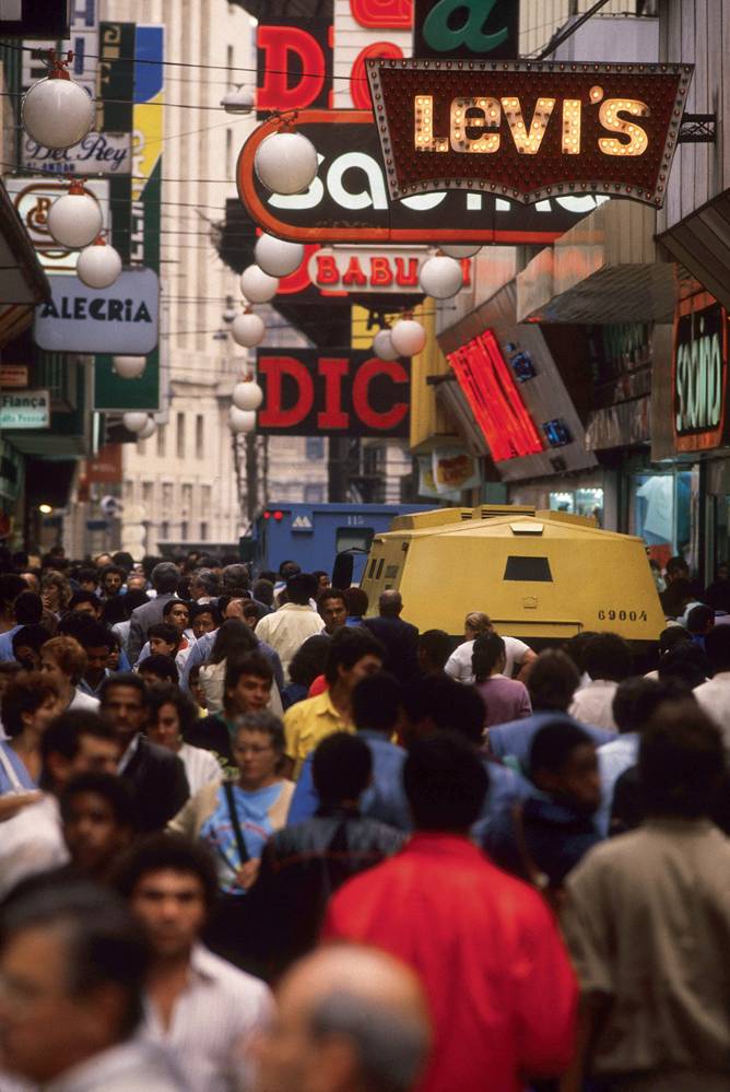 foto da rua direita, lotada de pessoas, com diversas placas de anúncio de marcas sobre suas cabeças