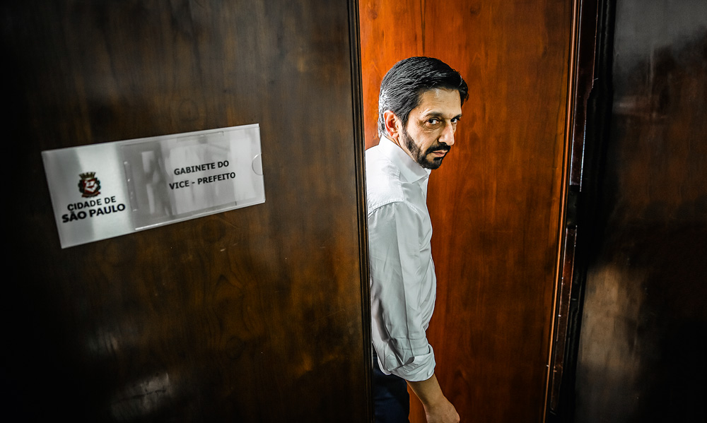 A imagem mostra Ricardo Nunes parado na porta de seu gabinete. Ele está a abrindo e há um pequeno letreiro escrito 