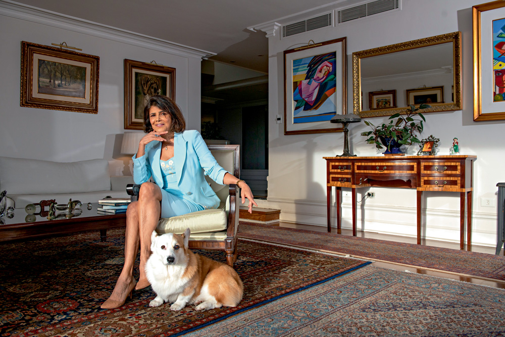 A astróloga Regina Braga com seu cãozinho Dexter posa de pernas cruzadas em cadeira no meio da sala de estar. O cachorro da raça Corgi está na sua frente, no chão, olhando para a câmera. Ela veste terninho e saia na cor azul-bebê e posa com uma das mãos no queixo. A sala tem quadros nas paredes, um grande tapete e aparador de madeira à direita.