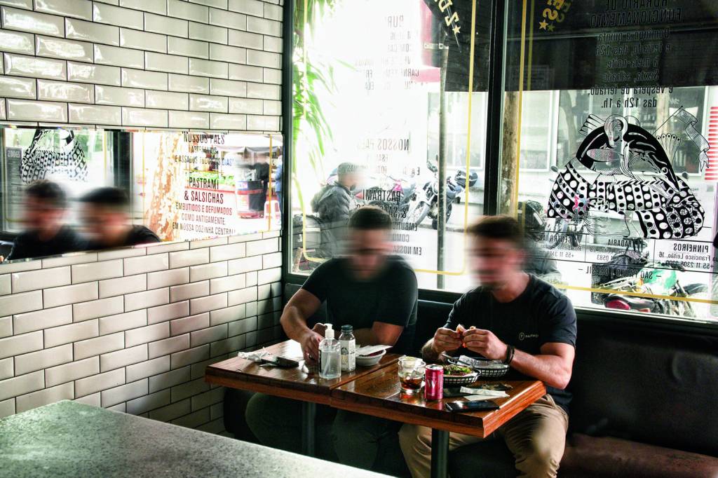 Dois homens sentados comendo em mesa de madeira ao centro da imagem, em frente a janelão de vidro que dá para a rua.
