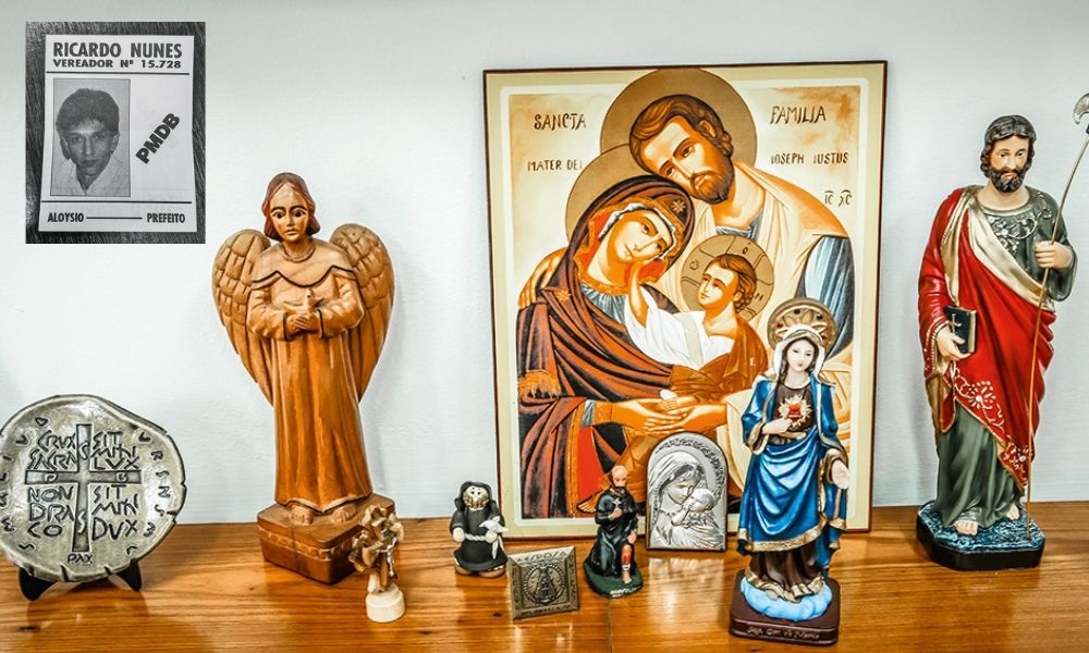 A imagem mostra figuras religiosas em uma mesa de madeira. Mais a cima, uma pequena foto de Nunes, ainda jovem, em uma cartaz escrito 