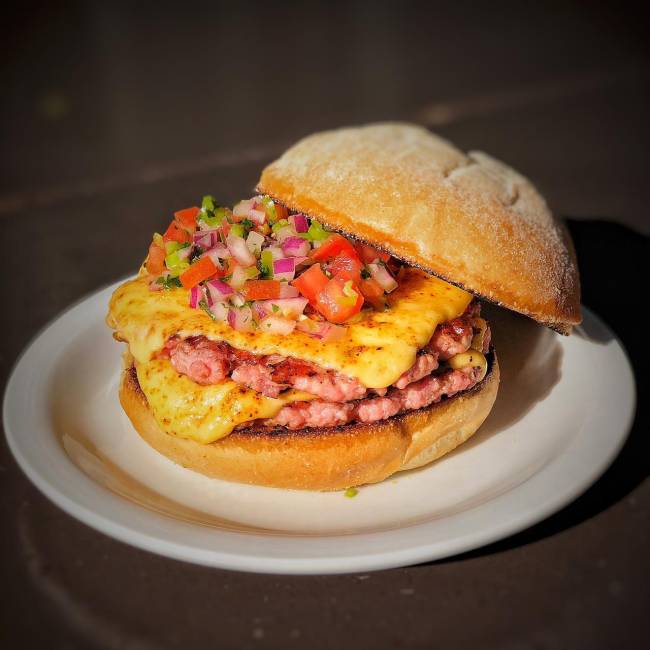 Hambúrguer sobre prato de louça branca em cima de mesa de madeira. Dois discos suínos (rosados) com queijo e vinagrete por cima, com o pão superior afastado para o lado.