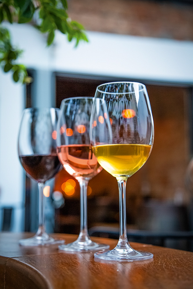 Três taças de vinho em sequência sobre tampo de madeira. Em primeiro plano vinho branco alaranjado, seguido por rosé e tinto.