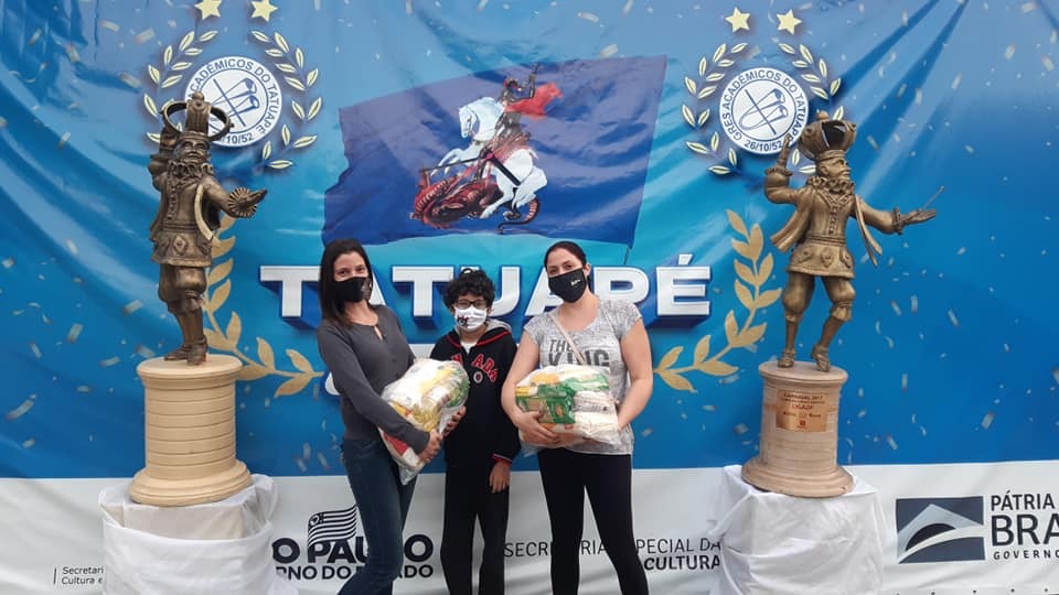 A imagem mostra três pessoas segurando sacos de arroz, todas de máscara. Ao fundo há um bandeirão com escudos e o nome da Acadêmicos da Tatuapé