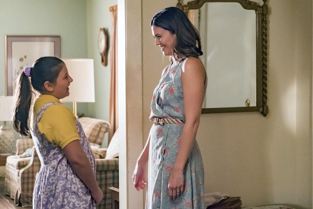 A imagem apresenta uma cena de This Is Us, em que a mãe Rebecca está de frente com a filha Kate em um quarto. As ambas estão sorrindo uma para a outra
