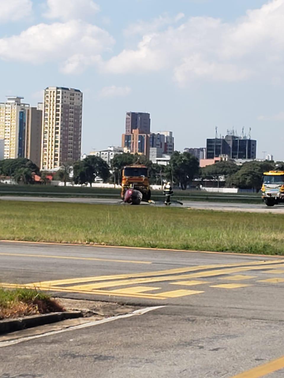 Imagem na altura de uma pessoa em pé. A distância, é possível ver o helicóptero tombado na pista do Campo de Marte.