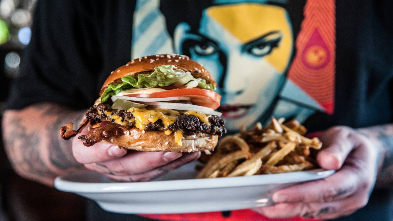 Foto horizontal com uma mão, à direita, segurando um hambúrguer em primeiro plano à direita. Ao fundo prato com batata frita.