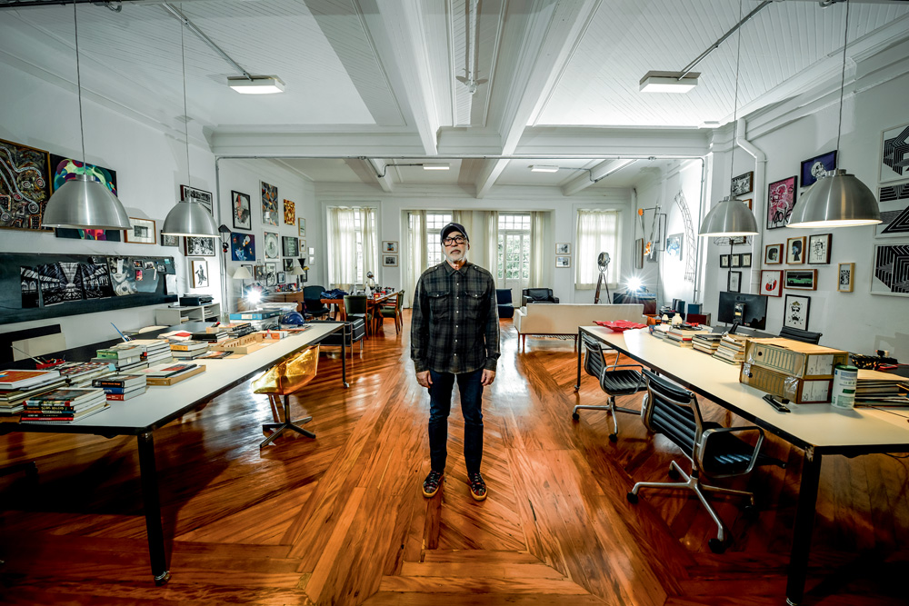 O cenógrafo Zé Carratu posa de pé no centro de seu estúdio, repleto de mesas, livros e janelões ao fundo. Veste camisa escura e óculos de grau.