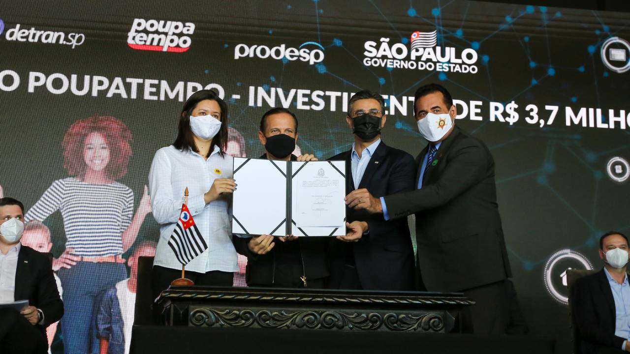 doria junto com o vice-governador e outras duas pessoas de pé, sorrindo para a foto, segurando documento assinado por doria
