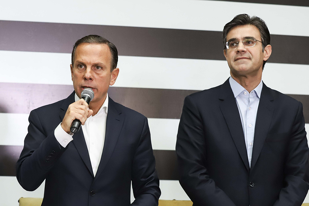 Imagem mostra João Doria em pé ao lado de Rodrigo Garcia, com bandeira do estado de SP ao fundo