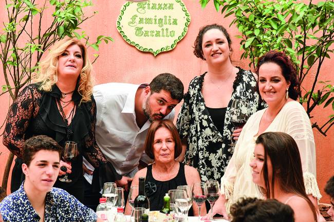 Família envolta de mesa de almoço com senhora matriarca ao centro e homem abaixado beijando o topo de sua cabeça.