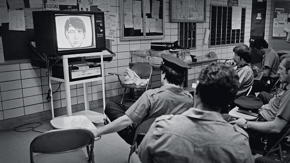 A imagem retrata uma sala de uma delegacia da década de 70. Preta e branca, é possível ver algumas pessoas sentadas em bancos simples em volta de uma televisão em que há um rosto projetado