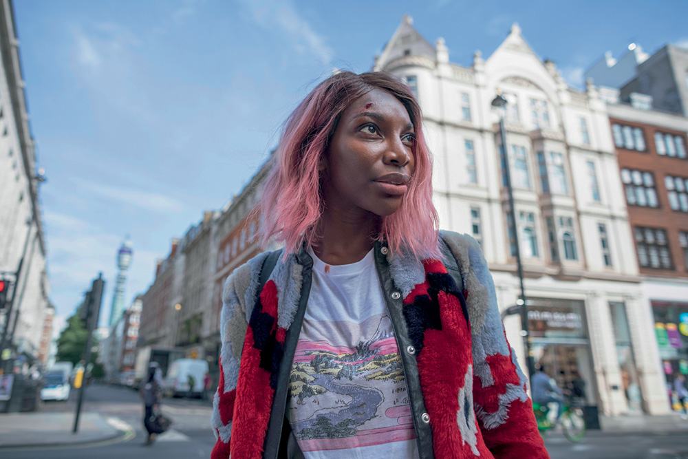 A imagem mostra Michaela na rua, com o cabelo rosa e olhando para o lado