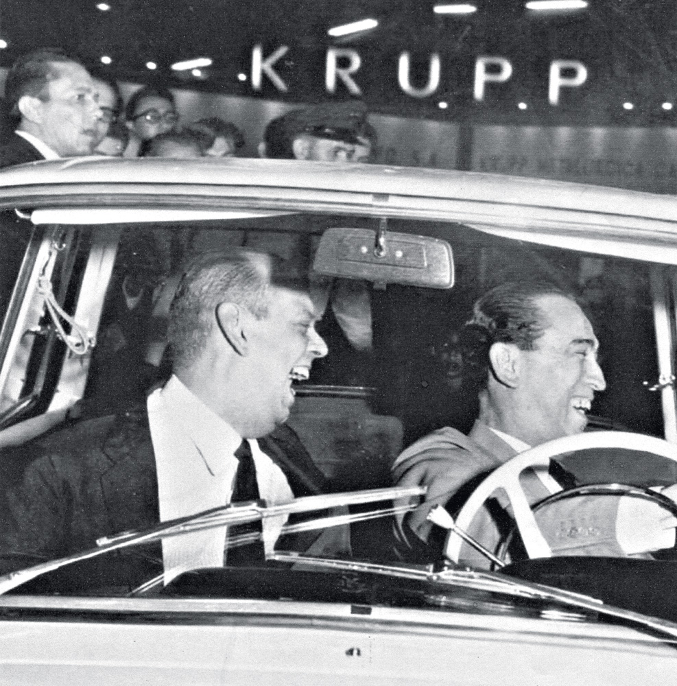 A imagem mostra Caio de Alcântara Machado e Juscelino Kubitschek dentro de um carro, com Juscelino no volante e os dois rindo ao olhar à direita.