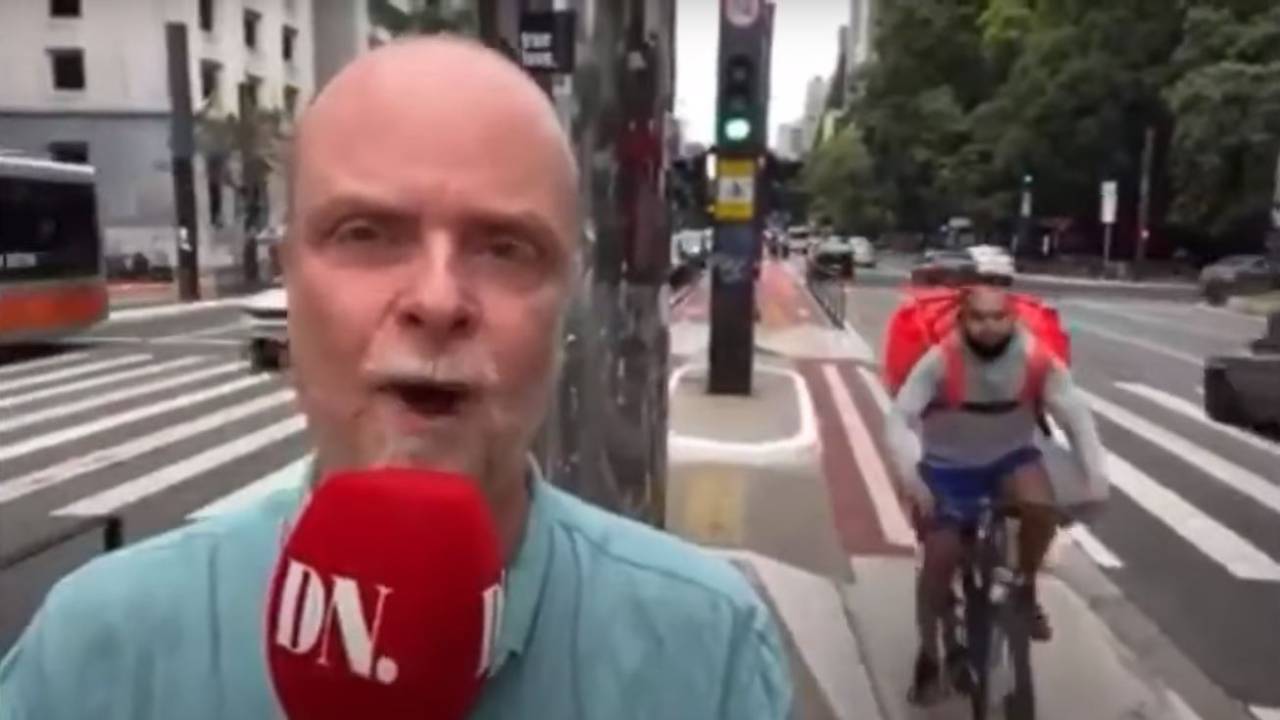 jornalista virado para a câmera com microfone na mão e ciclista vindo por trás dele