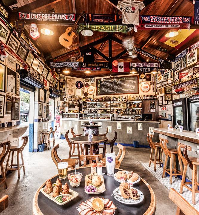 Salão do Bar do Luiz Fernandes com mesa posta com petiscos à frente em destaque, bancada ao fundo e banquetas em ambos os lados.