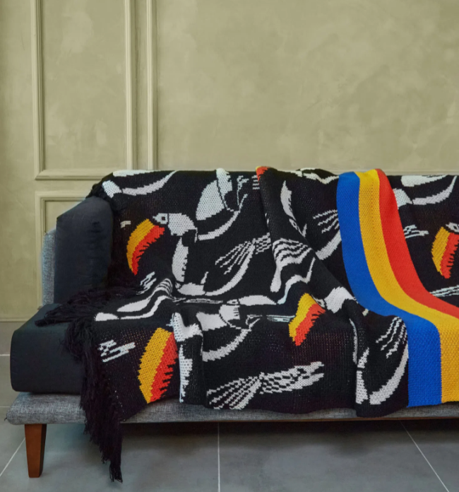 Um sofá tem manta estampada com vários tucanos. Fundo dela é preto