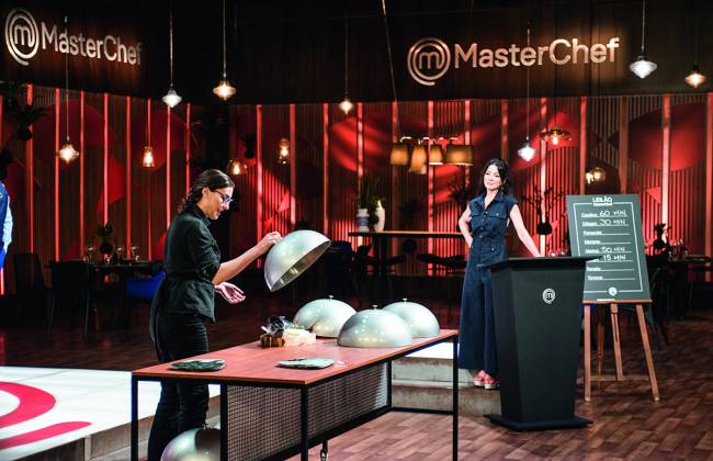Paola Carosella – MasterChef No estúdio do MasterChef. Mulher branca vestida de preto (Paola Carosella) à esquerda levantando forma esférica de prato sobre mesa de madeira. Ao fundo a apresentadora Ana Paula Padrão (mulher branca de cabelo preto) vestida de preto, encostada em púlpito do programa.