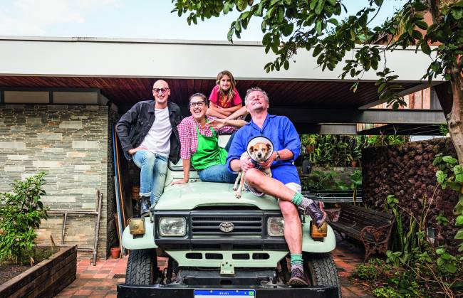 Família sobre jipe em frente à casa. Homem à esquerda, Paola Carosella ao centro, criança no topo e homem grisalho com cachorro no colo (à direita).