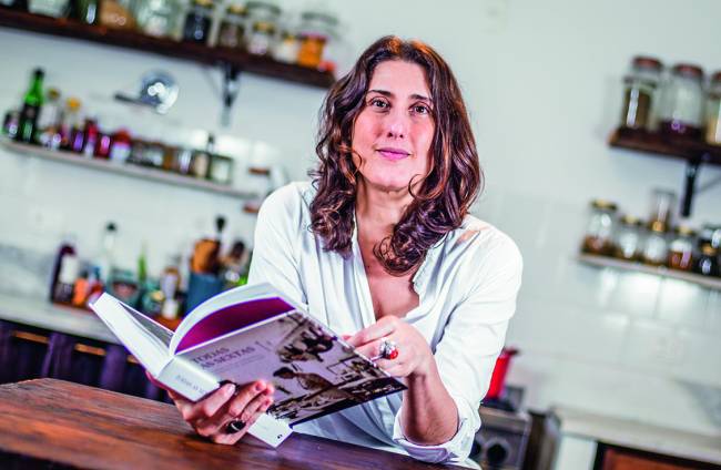 Paola Carosella (mulher branca de cabelo castanho) sentada atrás de bancada de madeira folheando livro.