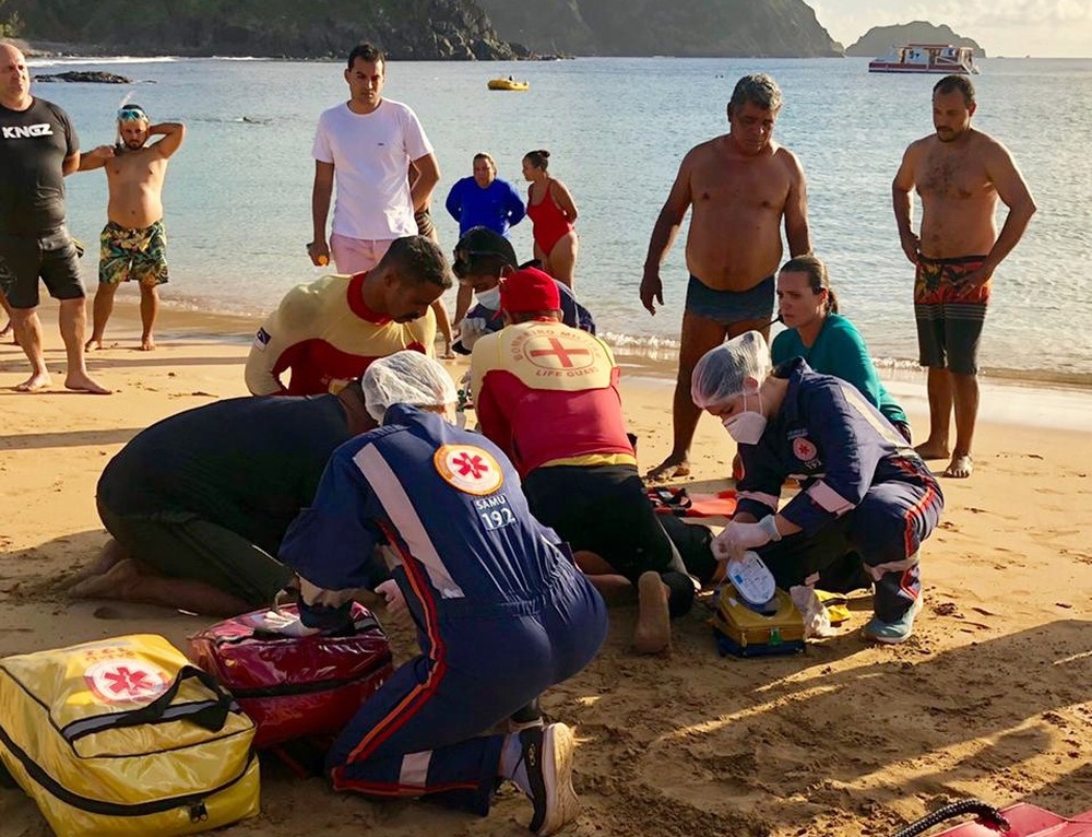 equipe de bombeiros e do samu na faixa de areia da praia debruçados sobre o corpo do turista, socorrendo-o
