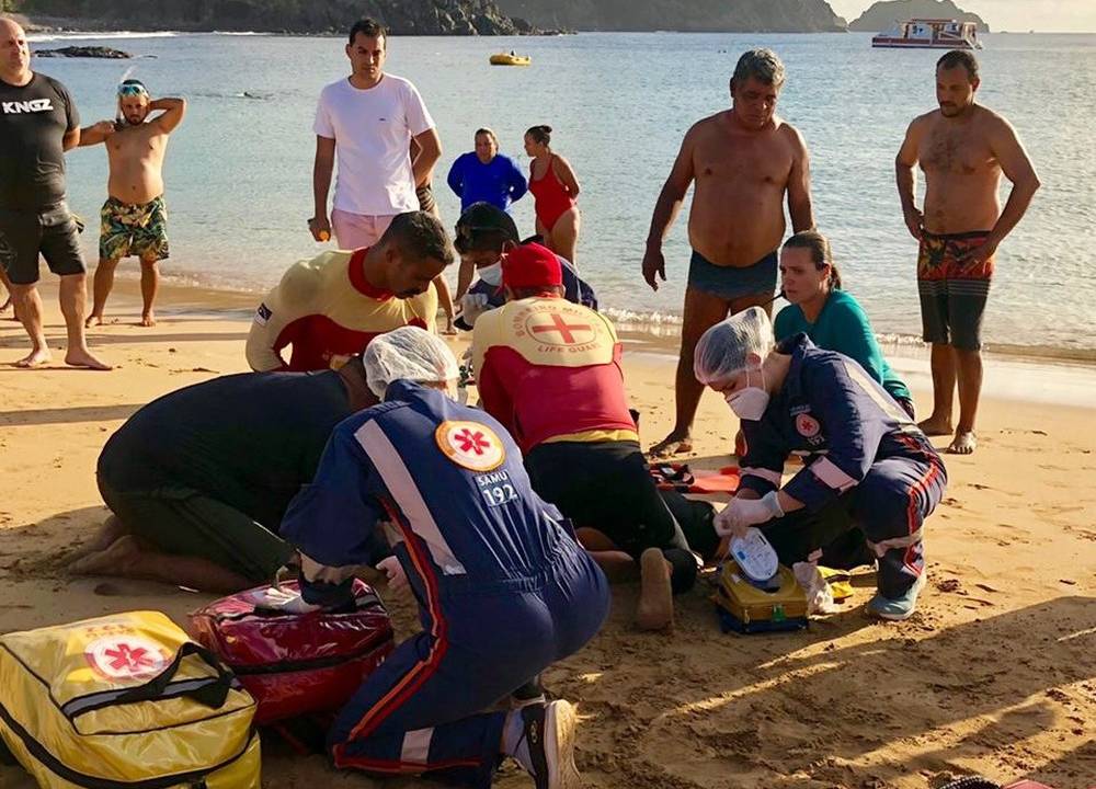 equipe de bombeiros e do samu na faixa de areia da praia debruçados sobre o corpo do turista, socorrendo-o