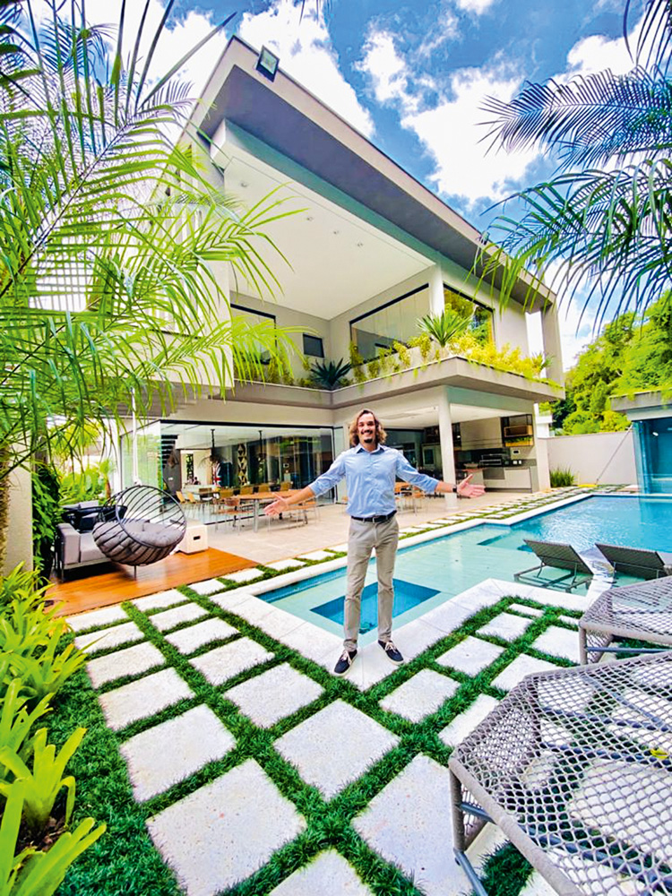super condominio litoral A imagem mostra Caio bem, pequeno, em frente à uma mansão enorme, de dois andares, árvores em volta e piscina.