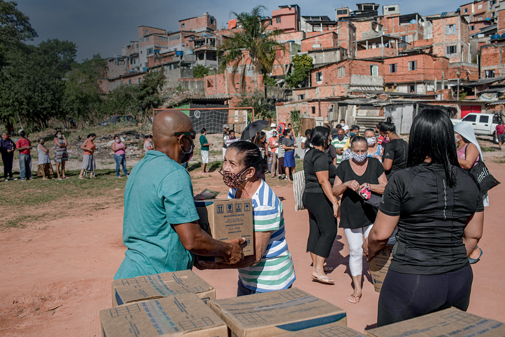 A imagem mostra uma fila de pessoas de máscara recebendo caixas de papelão. Ao fundo, há uma favela.