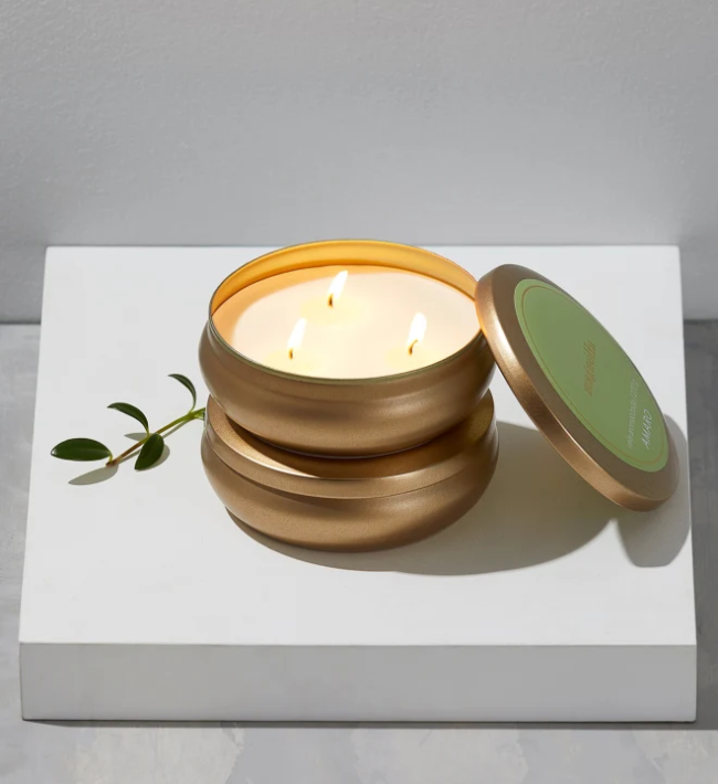 vela aromática Em uma caixa branca, uma vela com três pavios acesos em estrutura dourada. A tampa está apoiada ao lado e é verde