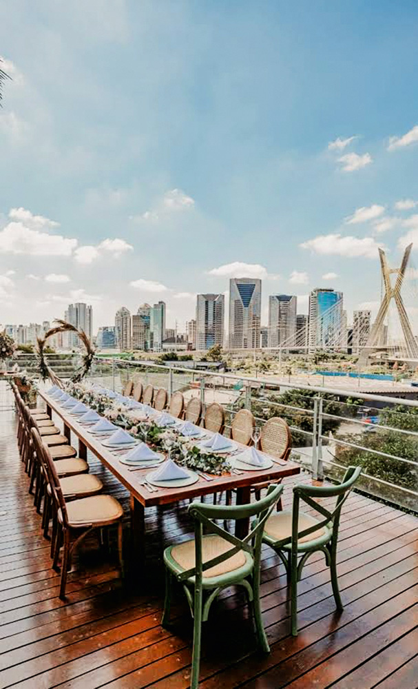 Foto mostra uma mesa formal, com muitos lugares, a céu aberto. Ao fundo, a cidade de São Paulo e prédios espelhados. Céu azul