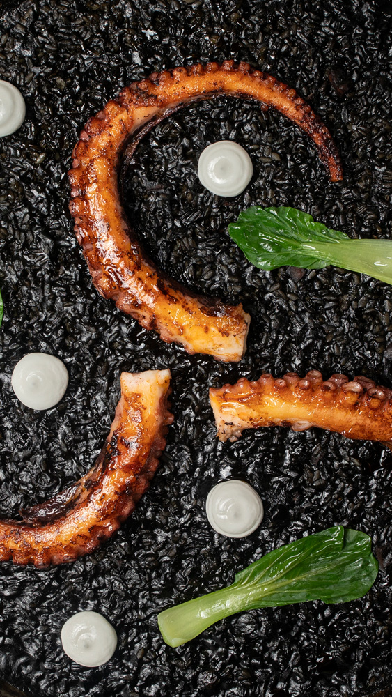 Foto aérea de arroz negro com tinta de lula e tentáculos de polvo grelhado espalhados por cima junto de bolinhas de aïoli.