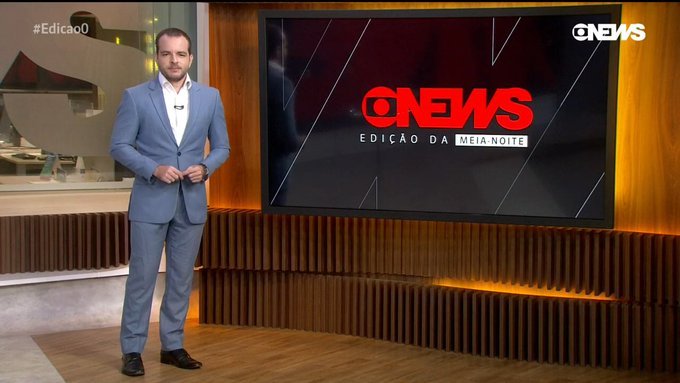 Imagem mostra Erick Bang em pé, usando terno, ao lado de telão que diz: "Globo News. Edição da Meia-Noite"