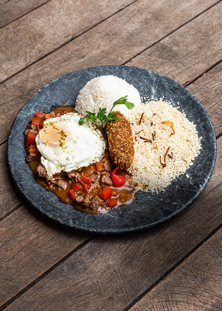 Prato de pedra sobre mesa de madeira com arroz, feijão, picadinho, ovo frito, bolinho alongado e farofa.