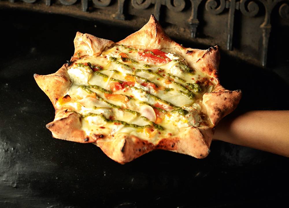 Pizza em formato de estrela da Napoli Centrale coberta por pesto em destaque no centro, sendo segurada por braço que vem da direita. Fundo escuro com arabescos metálicos.