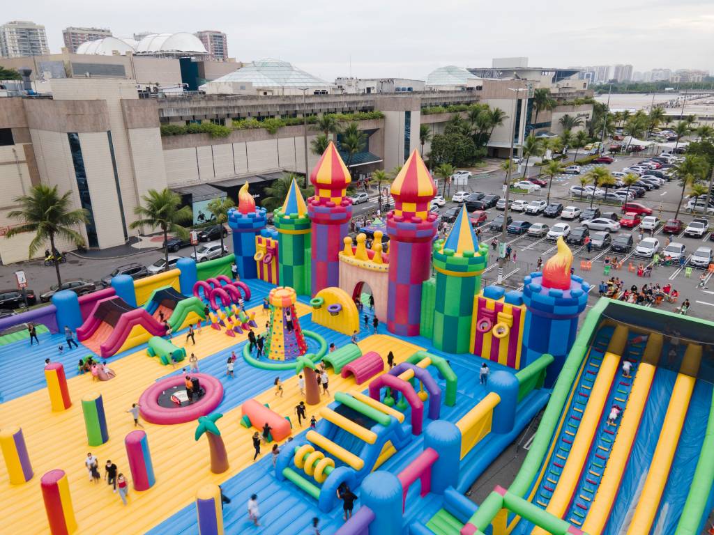 No estacionamento de um shopping, crianças brincam em uma estrutura inflável em formato de castelo colorida. Há carros em volta