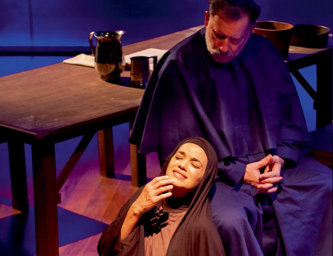 Em uma peça, uma monja tem expressão de tristeza, ajoelhada, enquanto um padre a observa