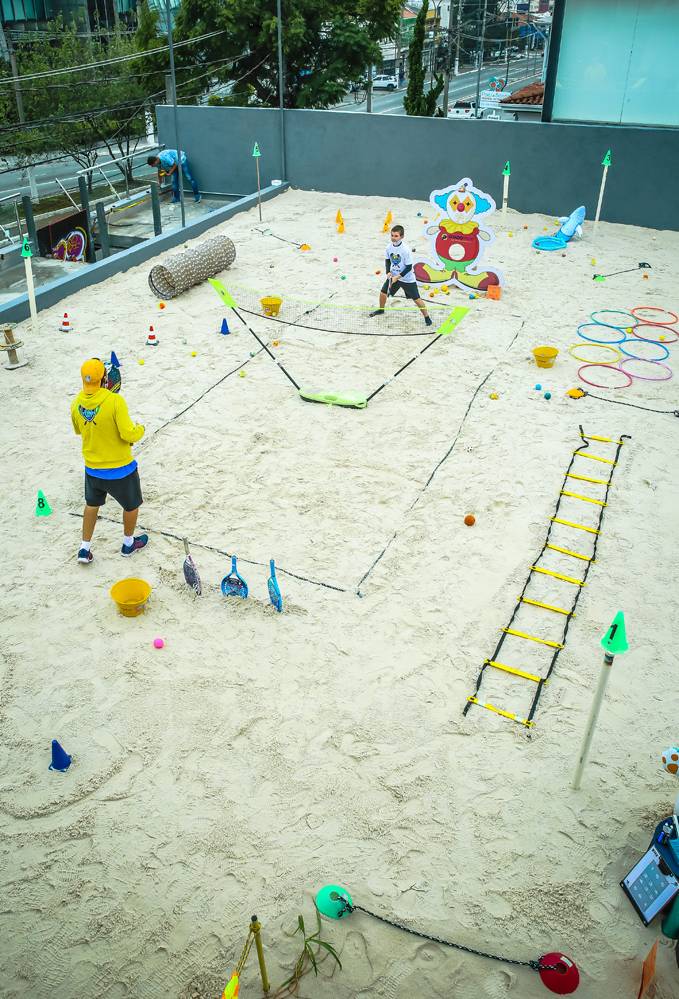 A imagem mostra Lalau com outra criança em uma quadra de areia. Há uma rede entre eles, vários objetos de treinos espalhados fora da quadra e a criança está em posição de expectativa com sua raquete