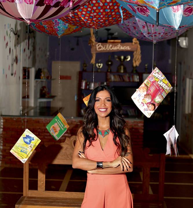 Amanda posa de braços cruzados com roupa na cor salmão. O cenário exibe guarda-chuvas coloridos no teto, de ponta cabeça.