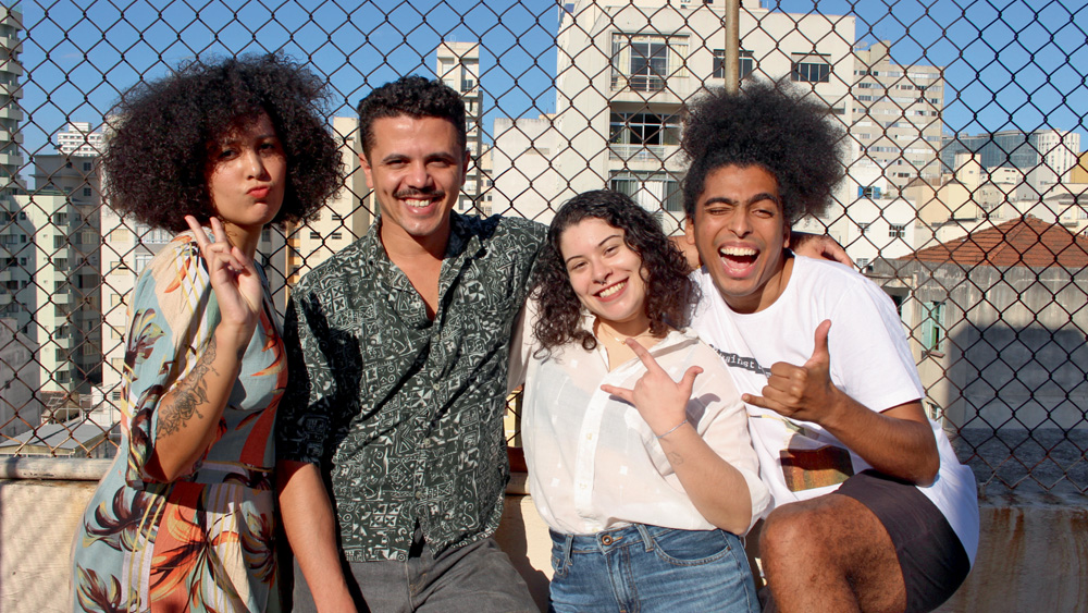 A partir da esquerda, Ana Beatriz Felicio, Vagner de Alencar, Gabriela Carvalho e Rômulo Cabrera aparecem abraçados em fente a uma grade de quadra. Em um local aberto e ensolarado, todos sorriem e acenam para a câmera.