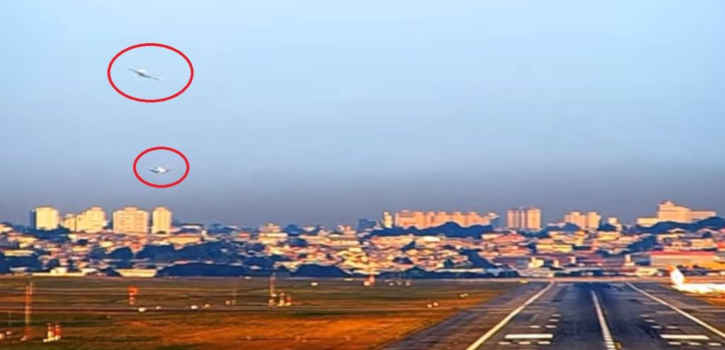 A imagem mostra o aeroporto de Guarulho, visto do chão. É possível ver duas aeronaves, circuladas de vermelho, uma fazendo uma curva no céu e outra pousando na pista.