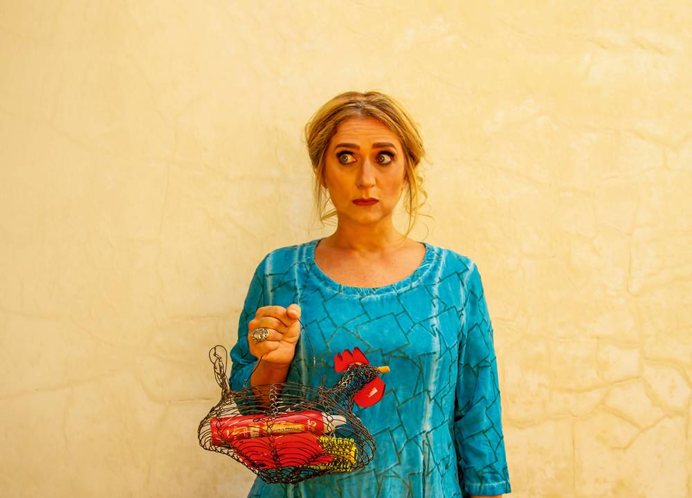 Mulher faz cara de espanto, de vestido azul, segurando uma cesta em formato de galinha