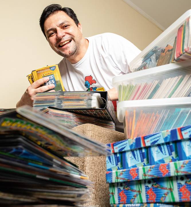 A imagem mostra Alvaro segurando alguns pacotinhos de figurinha em meio a uma pilha das pastas e caixas com seu acervo de álbuns e envelopes