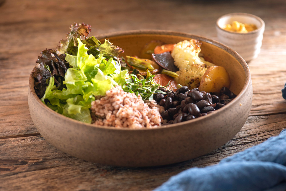 Cumbuca de cerâmica sobre mesa de madeira com arroz, feijão-carioquinha, vegetais salteados e salada verde mais farofa de castanhas.